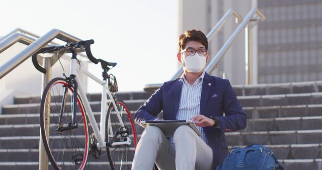 Businessman wearing formal attire and face mask sitting on steps. Bicycle parked beside him. He is holding and using a tablet while outdoors. Ideal for use in themes related to modern urban life, commuting, remote work, cycling, and health safety practices.