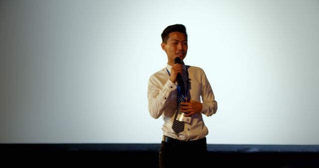 Young man in professional business attire is speaking confidently into a microphone on stage. Suitable for depicting public speaking, presentations, seminars, conferences, business events, and professional skills training.