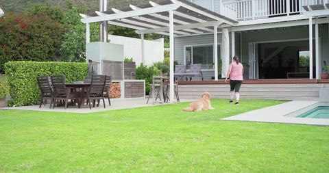 Mature woman walking toward deck with golden dog on lawn by pool and pergola dining