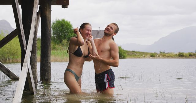 Young couple in swimwear standing in lake under wooden structure, sharing a playful and joyous moment. Ideal for depicting outdoor enjoyment, vacation vibes, summer activities, romantic moments, and carefree lifestyle.