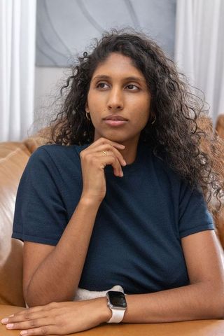 Young woman sitting on tan leather sofa thinking while touching chin, wearing navy top and smartwatc