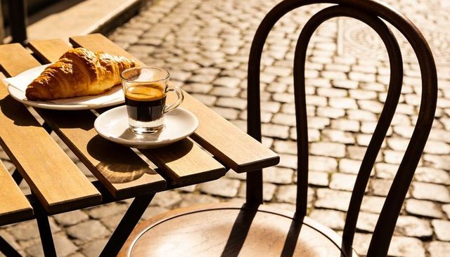 Sunlit espresso and croissant on bistro table casting chair shadows on cobblestone street
