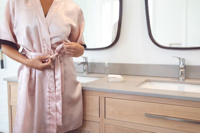 This image shows a woman tying the knot of her silk robe in a modern bathroom. The setting features a wooden vanity with two sinks and mirrors, suggesting a contemporary and luxurious home environment. This image can be used for articles or advertisements related to morning routines, self-care, home decor, or luxury lifestyle.