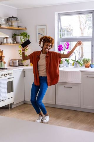 Joyful african american woman dancing in sunlit kitchen with headphones and smartphone