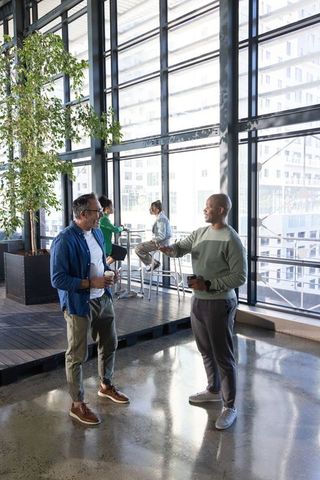 Diverse coworkers greeting and chatting while holding coffee in sunlit office atrium