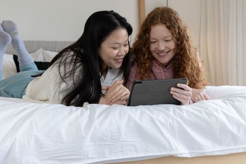 Two female friends lying on bed watching tablet, cozy bedroom bonding and streaming moment