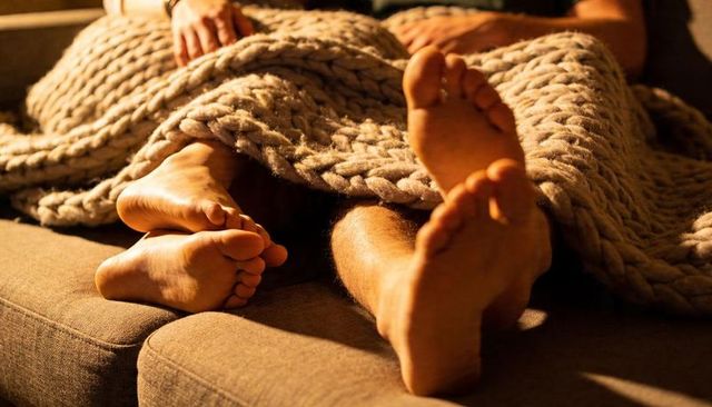 Couple relaxing on sofa under chunky knit throw, bare feet showing, warm ambient light