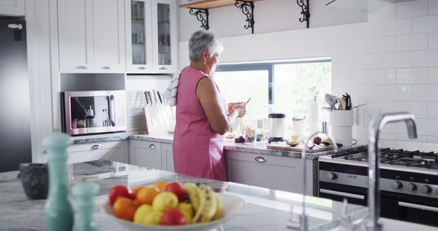 Senior woman preparing a healthy smoothie using fresh fruits in a modern, brightly lit kitchen. Shows active and healthy lifestyle, ideal for healthy living articles, senior well-being, diet and nutrition promotions, or advertisements for kitchen appliances and home decor.