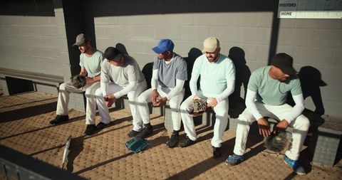 Sunlit baseball teammates sitting in dugout wearing caps and holding gloves before game