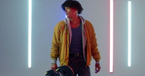 Urban youthful man holding skateboard with neon lights in studio