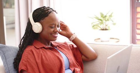 African woman relaxing on sofa wearing headphones using laptop, smiling