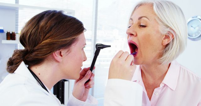 Health professional conducting thorough throat examination on senior woman using an otoscope. Senior woman's mouth is open while doctor examines. Useful for concepts related to healthcare, medical check-ups, elderly care, professional medical practice, diagnosis and treatment, and interaction between doctors and patients.