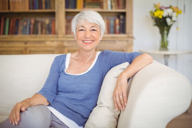 Senior woman with white hair smiling while sitting on a comfortable sofa in a cozy living room. Bookshelf and vase with flowers in the background add a homely touch. Ideal for use in lifestyle, retirement, and home comfort themes.