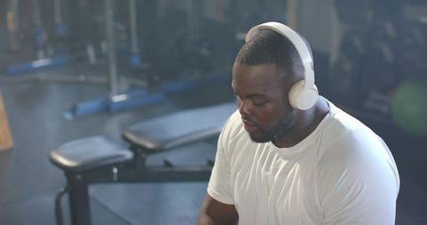 African american man wearing headphones resting on gym bench, focusing for training