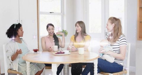 Sunlit moms chatting over coffee and bonding with baby around round wooden table
