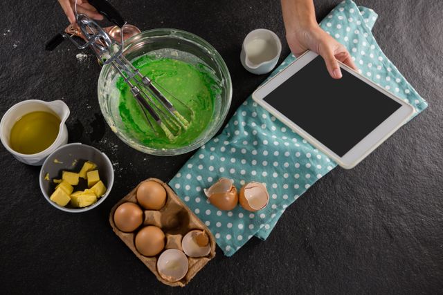 Woman using tablet while preparing batter in kitchen. Ideal for content related to cooking tutorials, digital recipes, culinary blogs, and home baking. Highlights use of technology in modern cooking.