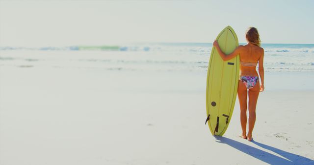 A young Caucasian woman walks towards the ocean carrying a surfboard, with copy space. Her relaxed posture and the tranquil beach setting suggest a leisurely day of surfing ahead.