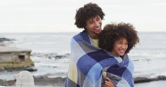 Couple warmly embracing wrapped in blue and white blanket by ocean, both smiling. Ideal for content related to love, travel, vacation, relaxation, family bonds, and advertising for outdoor activities or tourism.