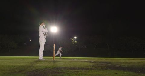 Floodlit cricket night fielder checking gloves beside stumps while bowler running on pitch