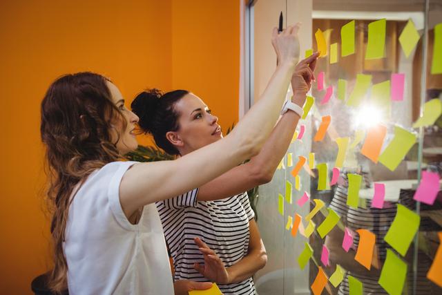 Two businesswomen are collaborating in an office environment, using colorful sticky notes on a glass wall to brainstorm and plan. This image is ideal for illustrating concepts related to teamwork, project management, creative thinking, and professional collaboration in a modern workspace.