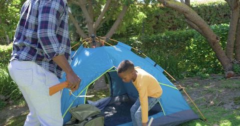 African american father and son pitching blue tent in sunlit backyard for family camping
