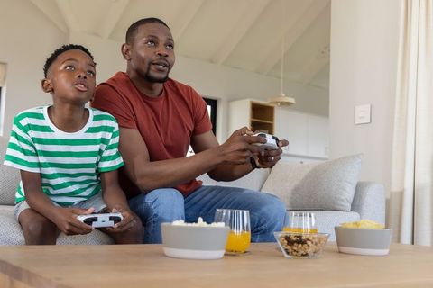 African american father and son playing console games on sofa, enjoying snacks and bonding