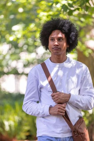 African american man standing in sunlit park holding leather crossbody bag strap