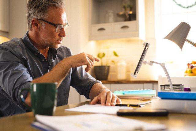 Middle-aged man wearing glasses having a video call on a tablet in a kitchen. He is pointing at the screen and appears engaged in the conversation. A coffee mug and various papers are on the table, suggesting a home office setup. Ideal for illustrating remote work, home office environments, and modern communication methods.