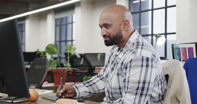 Biracial casual businessman sitting by desk using computer and holding pen in hand in office - Download Free Stock Photos Pikwizard.com