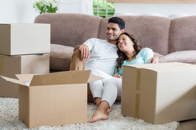 This image shows a happy couple sitting on the floor in their living room, surrounded by moving boxes. They are smiling and appear relaxed, suggesting they have recently moved into a new home. This image can be used for themes related to moving, new beginnings, home life, relationships, and domestic happiness.