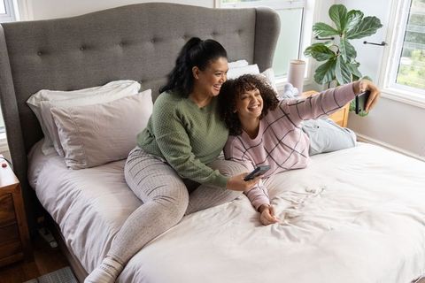 Two diverse women in cozy loungewear laughing and taking selfie on bed with plant