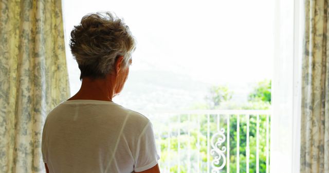 An elderly woman with short grey hair is standing and looking out a window with a scenic view outside. The curtains are open, revealing bright daylight and greenery. This can be used to depict themes of solitude, contemplation, retirement, peaceful living, or reflecting on nature.