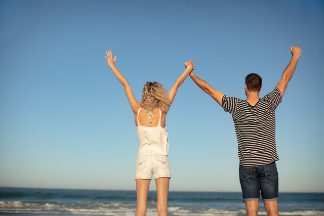 Rear view of couple standing together with arms up on the beach