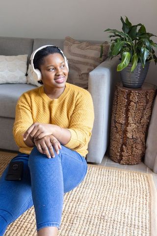 African american woman sitting on rug with headphones, cozy living room relaxation and indoor plant