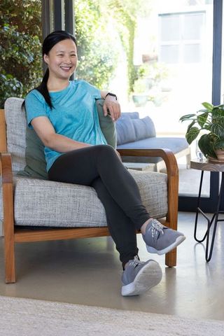Mature asian woman sitting, smiling in modern living room wearing athleisure and smartwatch