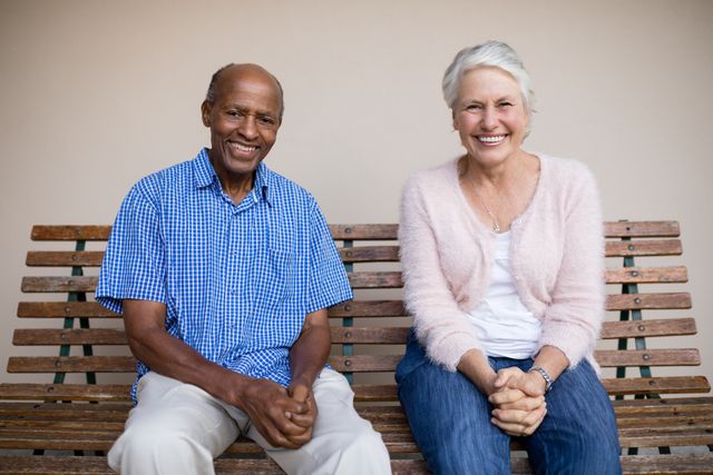 Portrait of smiling senior woman and man sitting on bench against wall at retirement home