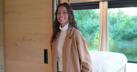 Smiling woman wearing tan coat by bed in modern minimalist bedroom with greenery