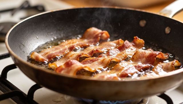 Sizzling bacon strips frying in nonstick skillet with bubbling fat and steam on gas stove