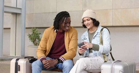 Young female and African American male friends checking travel plans on phone while sitting with lug