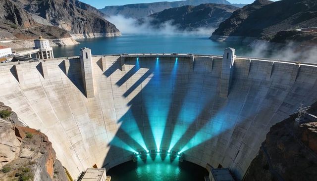 Curved concrete dam emitting turquoise water jets and light beams over canyon reservoir