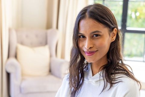 Smiling african woman sitting in cozy living room wearing white hoodie by armchair in natural light