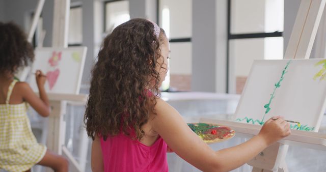 Young girls with curly hair engaging in painting activity on canvases set up on easels. Ideal for educational materials, promotional content for art schools, and blogs about children's activities and creativity.