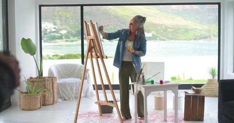 Senior woman painting in sunlit lakeside studio with easel and canvas, creative hobby
