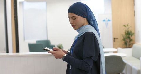 Nurse wearing two-tone headscarf holding smartphone at clinic reception checking messages