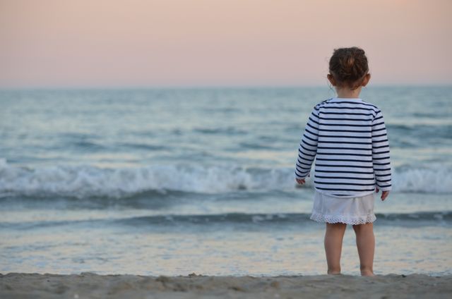 Young girl with a striped shirt stands on sandy beach watching waves under a twilight sky. Use for themes of childhood, innocence, relaxation, nature, family vacations, and coastal living. Perfect for advertising summer travel destinations, parenting blogs, or inspirational content.