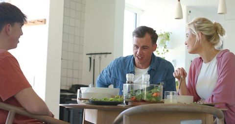 Family sharing casual mealtime conversation around sunlit kitchen table with fresh salad