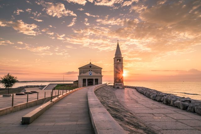 View of coastal church and bell tower in Caorle, Italy during sunrise. Tranquil scenery featuring historical architecture, open pathways, and vibrant sky, ideal for travel guides, tourism promotions, and inspirational backgrounds.