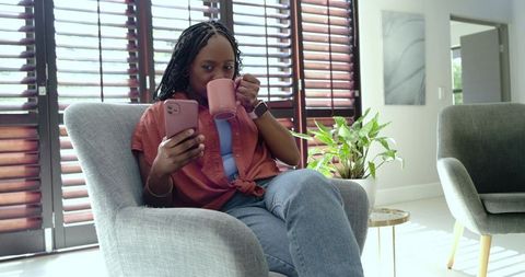 African woman sipping coffee, checking smartphone in cozy modern living room