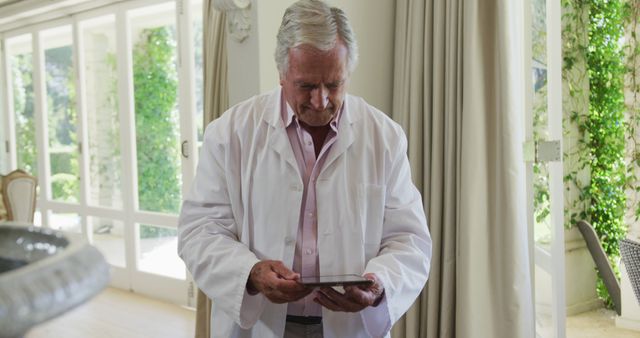 Senior man standing indoors wearing a white coat, looking at a tablet. Appears to be a medical professional, perhaps a retired doctor, using technology at home. Scene might be set in a bright and spacious home with plant background. Useful for portraying themes of technology use among seniors, medical professionals in a relaxed setting, or elderly people staying updated.