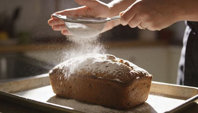 Baker sifting powdered sugar over sunlit loaf on metal tray, hands dusting with sieve